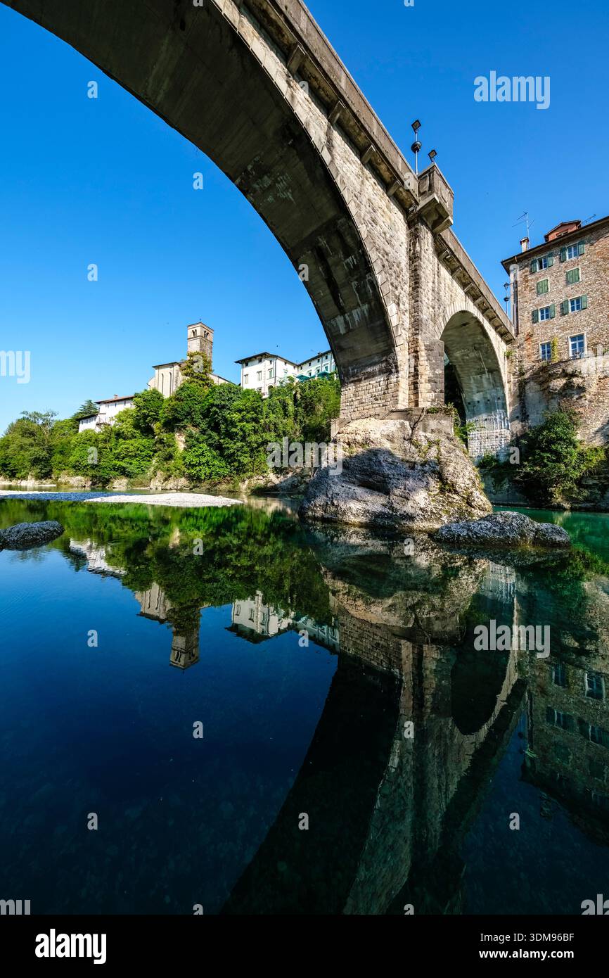 Die Steinbrücke Devil's Bridge überspannt den türkisfarbenen Natisone River mit der Kirche San Francesco und alten Häusern in der Ferne. Stockfoto