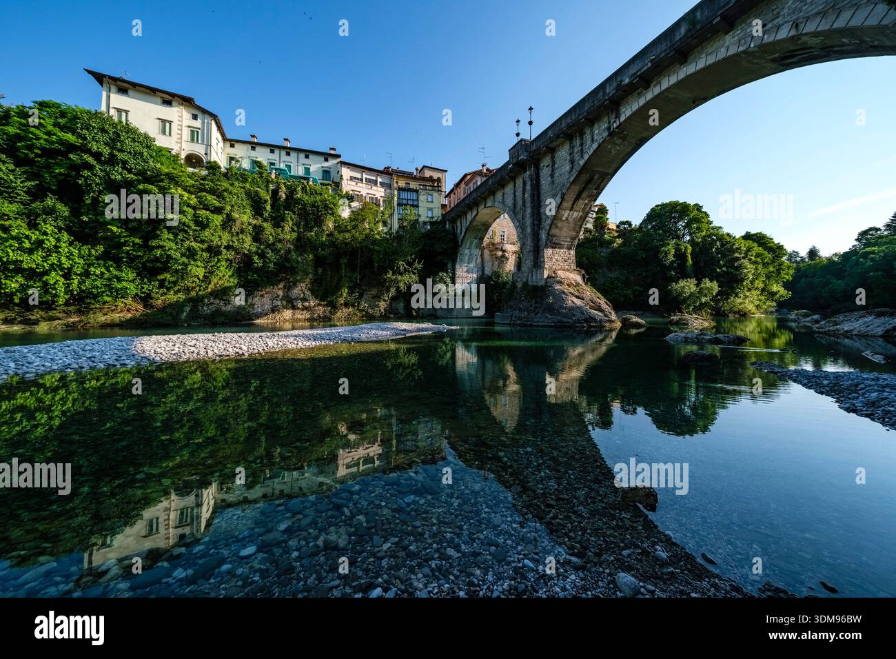 Die Steinbrücke Devil's Bridge überspannt den türkisfarbenen Natisone River mit historischen Bürgerhäusern von Cividale del Friuli in der Ferne. Stockfoto