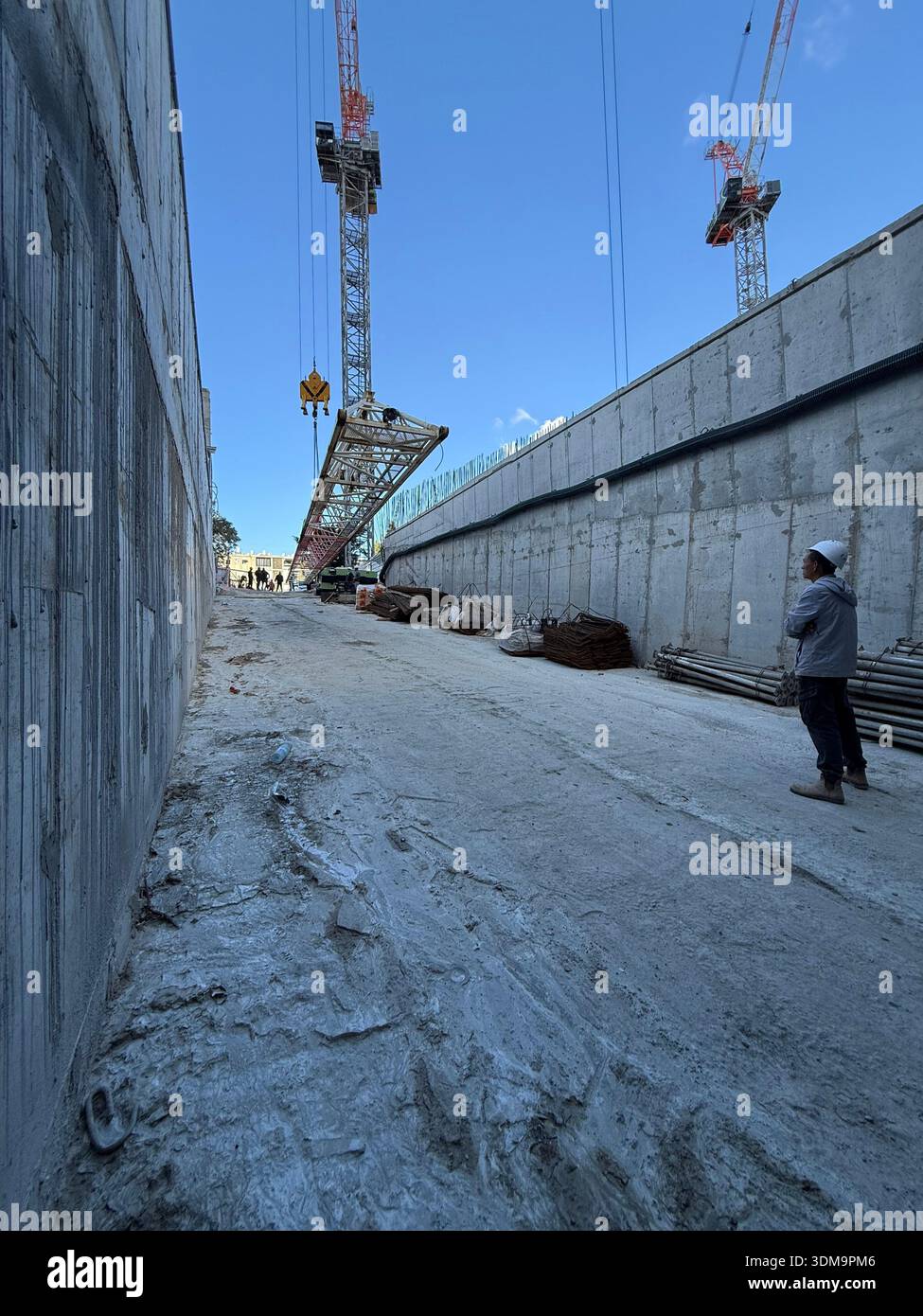 Ein Bauarbeiter beobachtet, wie ein Turmkran einen großen Auslegerabschnitt unter klarem Himmel in eine tiefe Betongründung senkt. - Smartphone-aufgenommenes Stockfoto