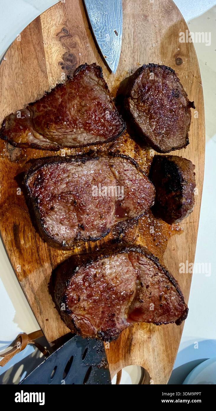 Perfekt gebratene, saftige mittelseltene Steaks ruhen auf einem rustikalen Holzschneidebrett mit einem scharfen Kochmesser. Eine köstliche, hochwertige kulinarische Szene. - Smartphone-aufgenommenes Stockfoto