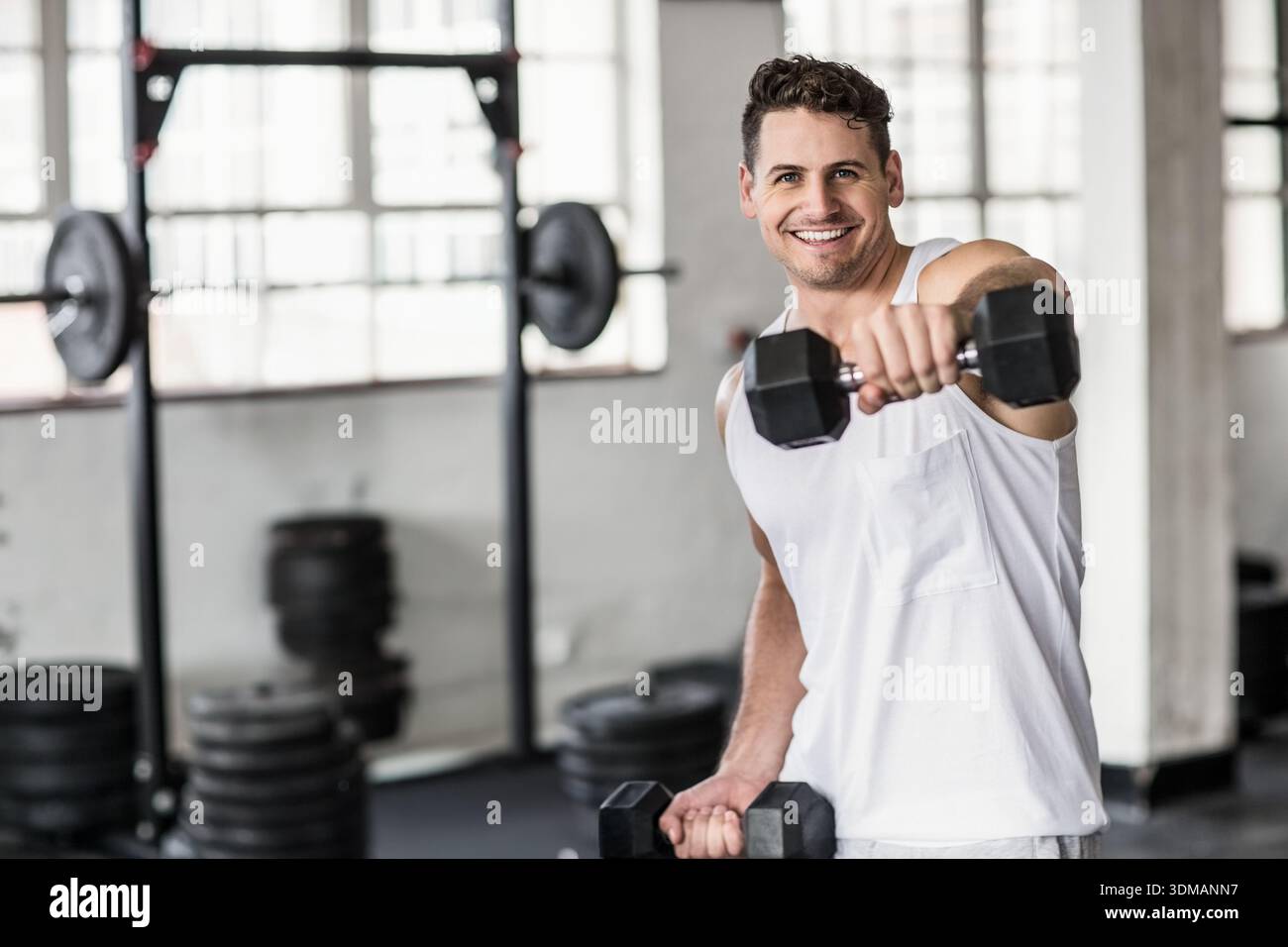 Mann in den Zwanzigern, der Tanktop trägt, im Fitnessstudio vorne hochfährt, Hexenhantel hält, Kopierraum. Fitness, Workout, sportlich, Gewichte, Langhantel, Rack, Stockfoto