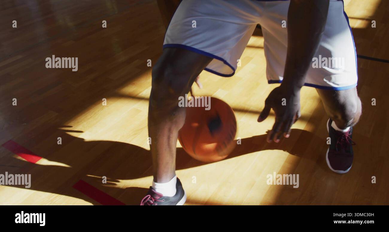 Dribbling reifer Spieler, der Basketball auf der roten Linie des Fitnessstudios aus Holz bewegt, in weißen Shorts und dunklen Schuhen Stockfoto