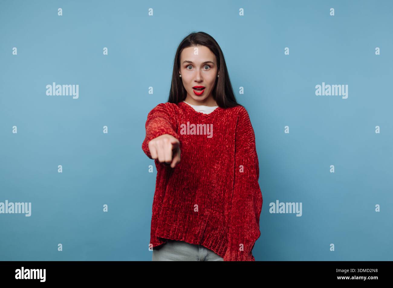 Eine junge Frau steht in einem Atelier mit blauem Hintergrund. Sie hat lange braune Haare und trägt einen roten Pullover. Sie zeigt mit einem Ex direkt auf die Kamera Stockfoto