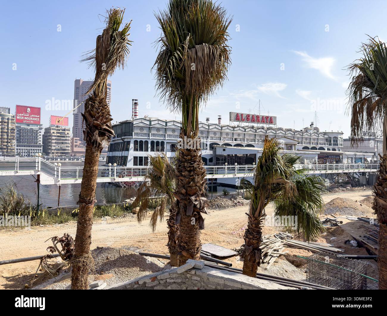 Blick auf den Fluss entlang der Nil Corniche in Kairo, Ägypten, mit Palmen, städtischer Infrastruktur am Wasser und einem schwimmenden Restaurant am Nil R Stockfoto
