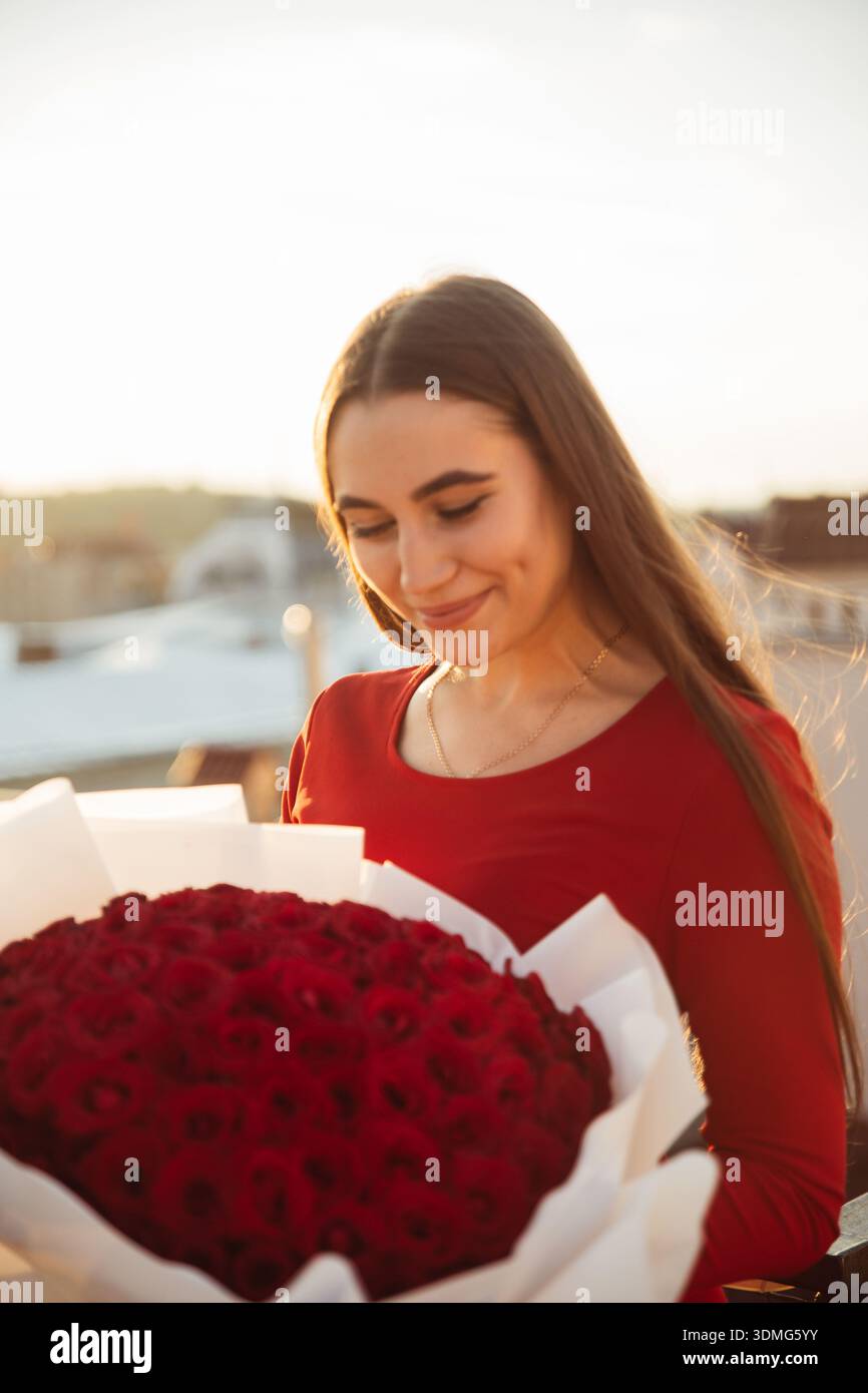 Eine Frau steht auf einem Dach und lächelt, während sie einen großen Strauß roter Rosen hält. Die Sonne geht im Hintergrund unter. Stockfoto