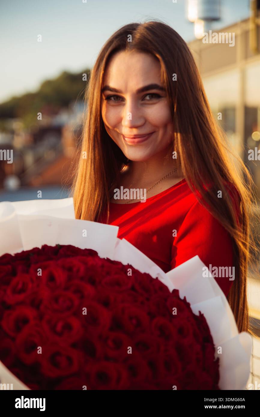 Eine Frau steht draußen und hält einen großen Strauß roter Rosen. Sie lächelt in die Kamera, während das Sonnenlicht auf ihr Haar scheint. Stockfoto
