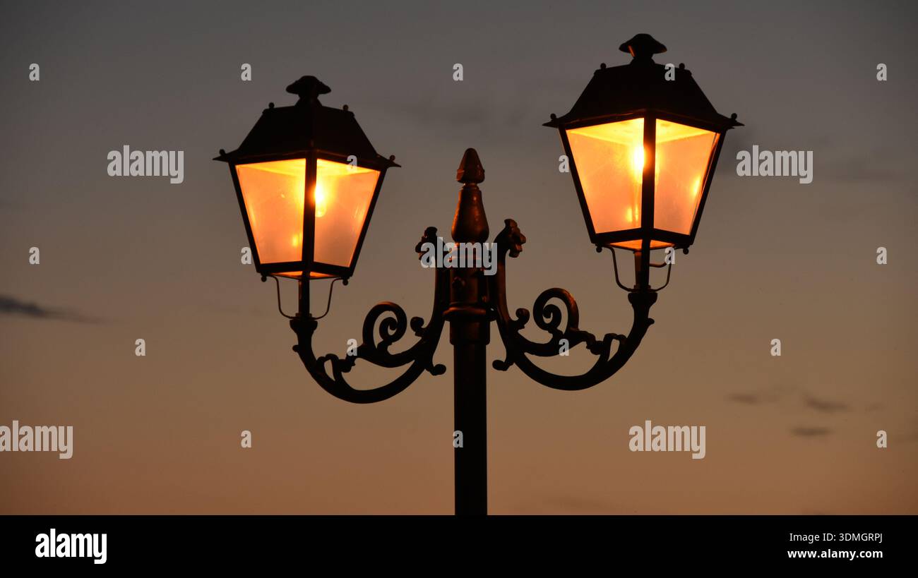 Eine Straßenlaterne im antiken Stil mit zwei Laternen leuchtet mit warmem orangenem Licht vor einem dunkleren Abendhimmel in der Abenddämmerung und schafft so eine romantische und nostalgische Atmosphäre Stockfoto