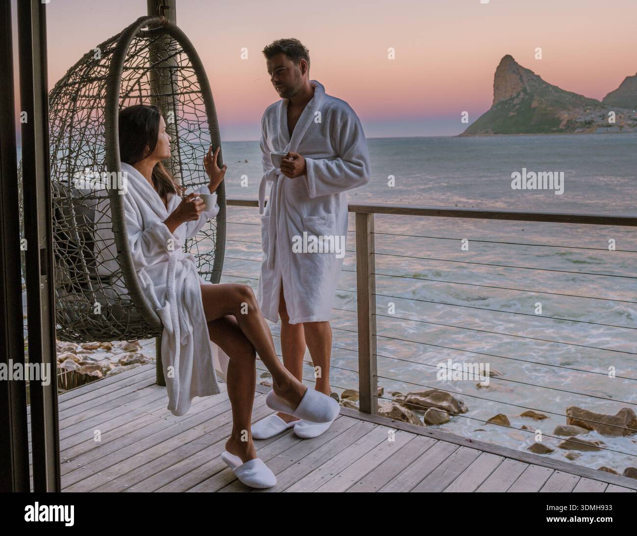 Ein Paar in gemütlichen Bademänteln genießt dampfende Tassen Kaffee, während er den atemberaubenden Sonnenaufgang über dem Meer in Kapstadt, Südafrika, bestaunt. Stockfoto