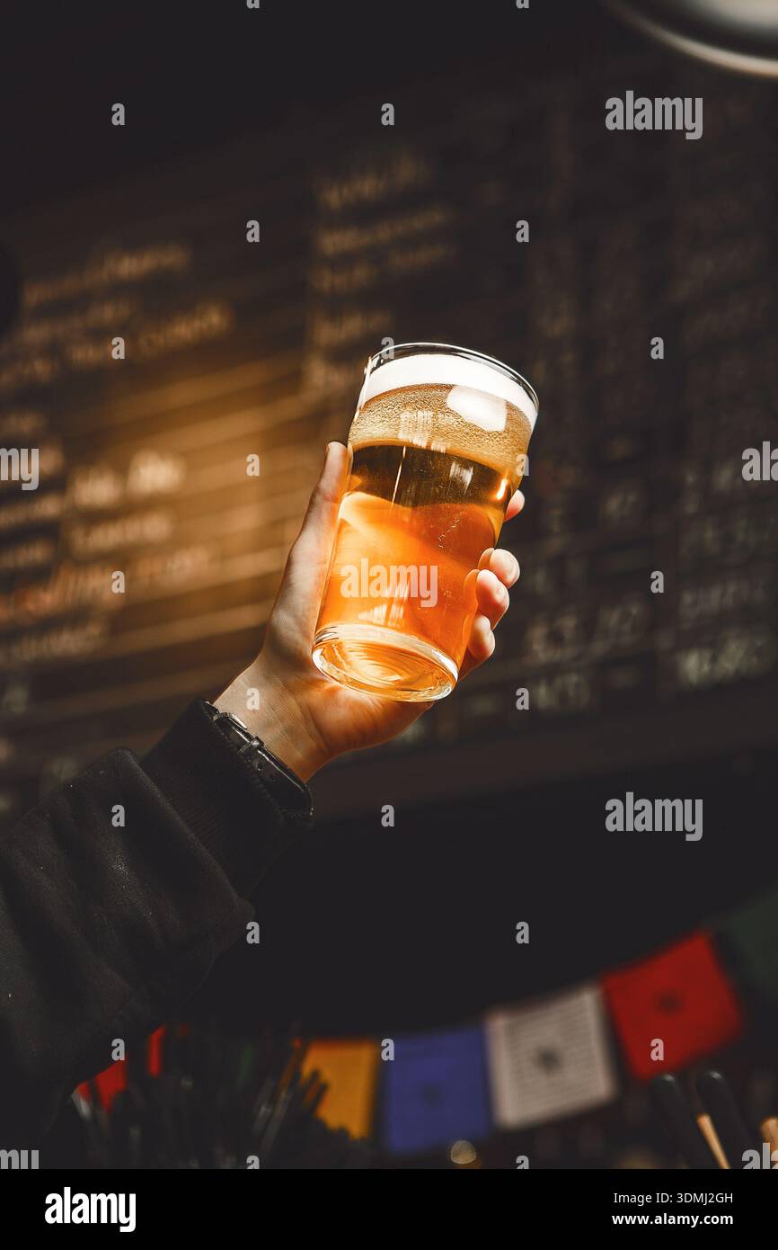Hand hält erfrischendes Bier Glas in lebendiger Bar-Atmosphäre mit farbenfroher Einrichtung Stockfoto