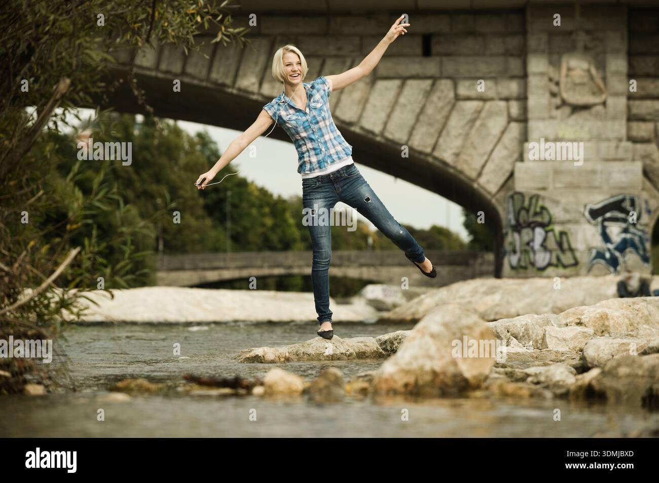 Eine verspielte junge Frau, die auf Steinen an einem flachen Fluss in der Stadt balanciert Stockfoto