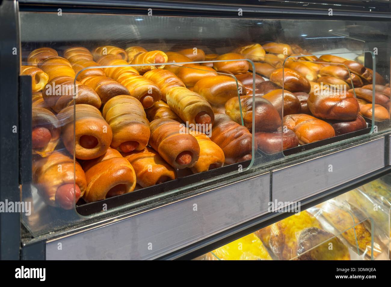 Reihen von herzhaften Hot Dogs in weichen, goldenen Teig gewickelt, zusammen mit verschiedenen süßen Brötchen, die eine verlockende Auswahl an Backwaren zum Verkauf in einem Vibr präsentieren Stockfoto