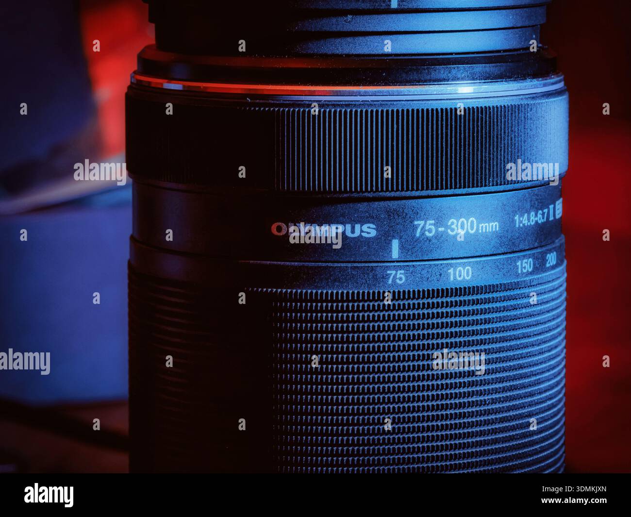 Nahaufnahme eines Olympus 75–300 mm Teleobjektivs mit Fokus- und Zoomringen unter dramatischer roter und blauer Studiobeleuchtung. Stockfoto