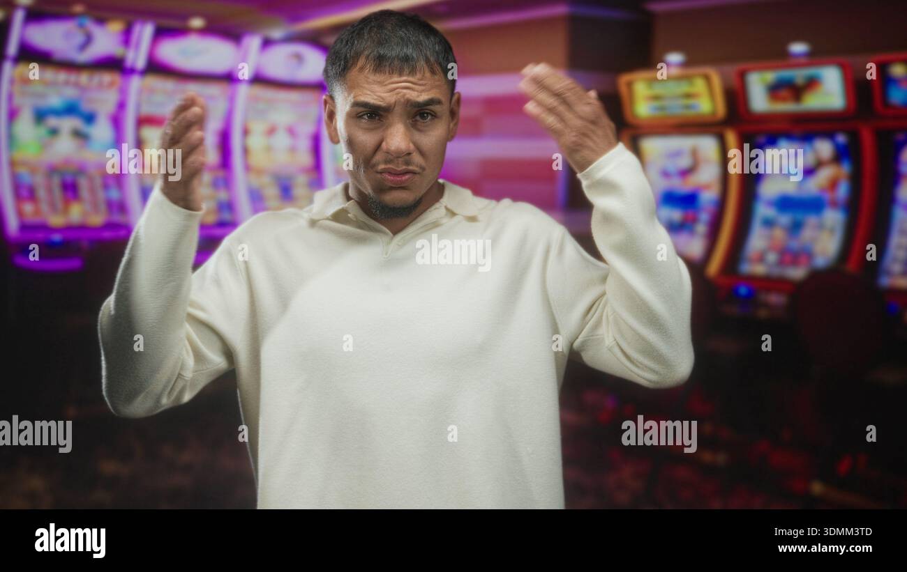 Mann, der die Brust mit der rechten Hand berührt, mit weißem Pullover, vor beleuchteten Spielautomaten in einem Casino stehend, mit einem angespannten Gesichtsausdruck Stockfoto
