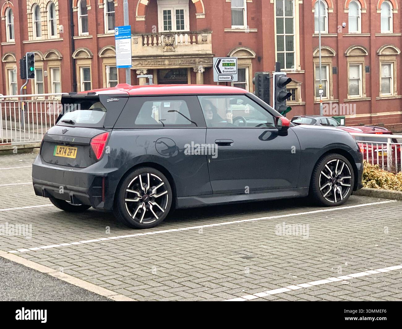 Graues Fließheck mit kontrastierendem rotem Dach, das auf einem britischen Parkplatz mit historischer roter Backsteinarchitektur im Hintergrund geparkt wird Min. Graues Auto-rotes Dach - Smartphone-aufgenommenes Stockfoto