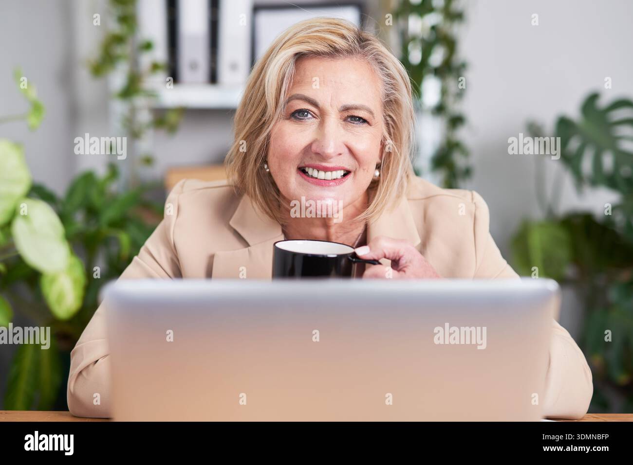 Freundliche Geschäftsfrau genießt Kaffee am Schreibtisch mit Laptop im hellen, modernen Büro Stockfoto