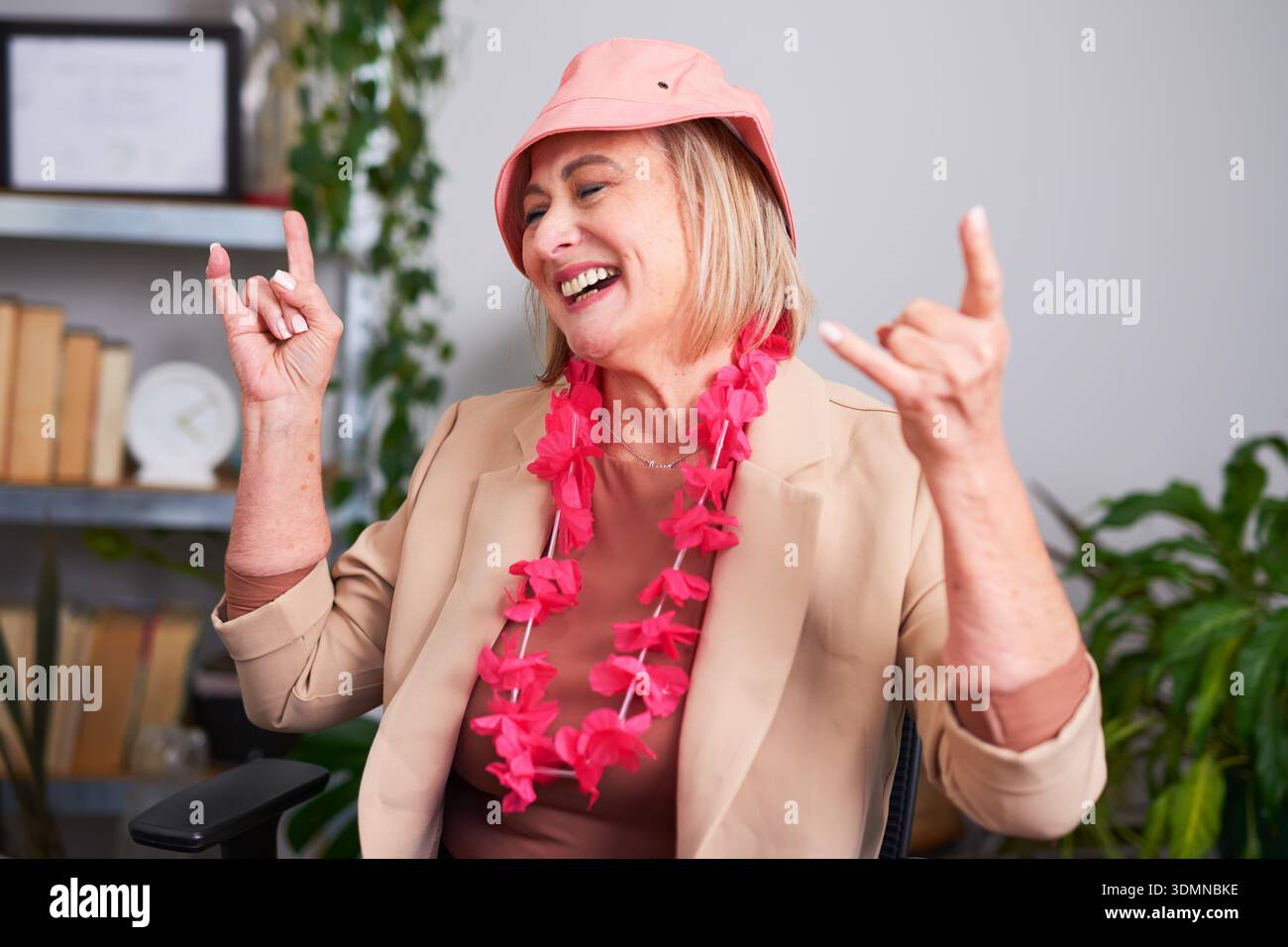 Fröhliche Seniorin feiert bei der Arbeit mit Pink hat und Floral Lei im Büro Stockfoto