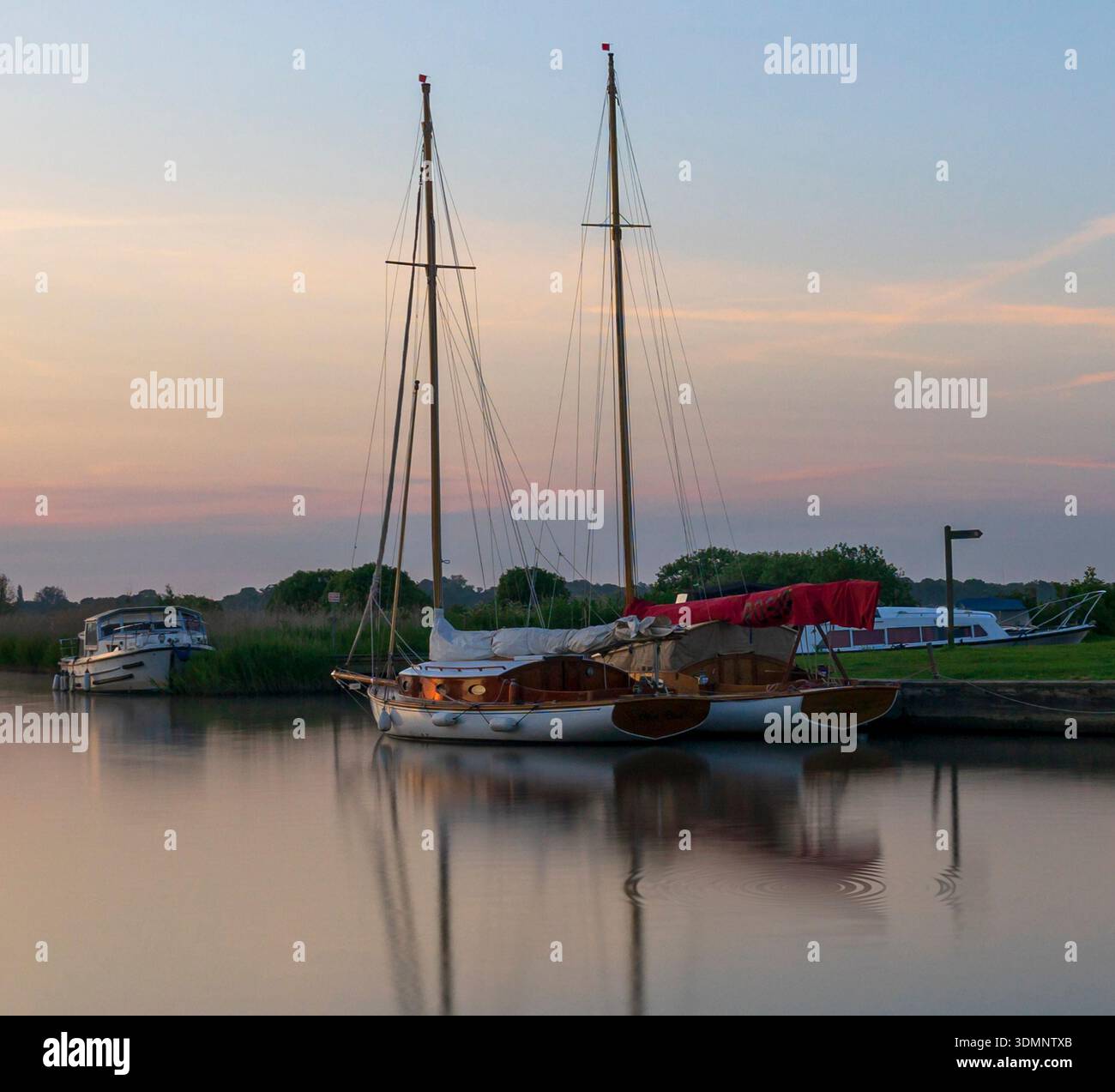 Thurne Mill Dyke, Norfolk Broads, Großbritannien – Sonnenlicht über festgefahrenen Booten mit ruhigen Wasserreflexionen, die durch lange Belichtung und malerische Darstellung verstärkt werden. Stockfoto