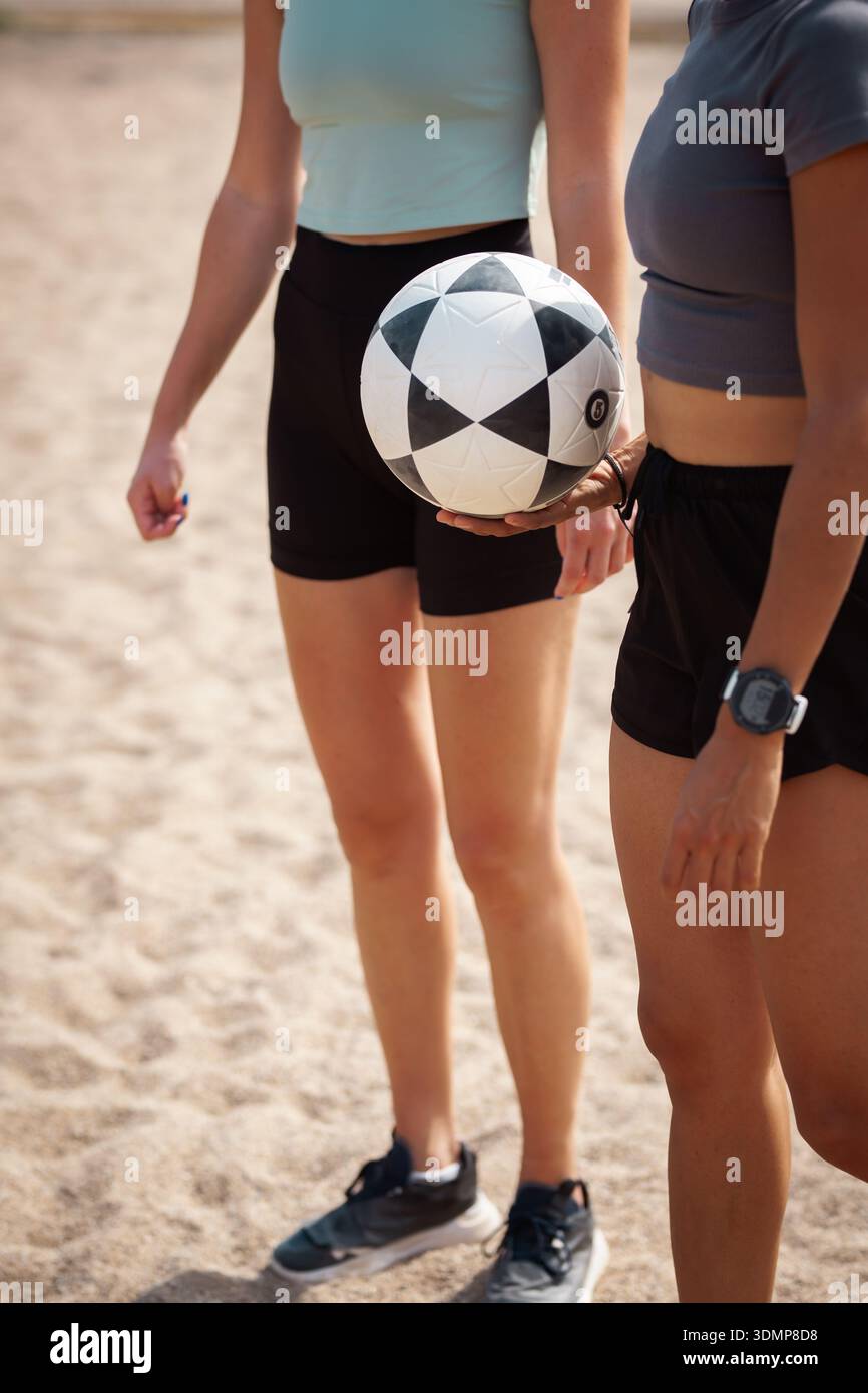 Fitnessbegeisterte halten einen schwarz-weißen Ball am Strand, während sie sich auf ein lustiges Spiel in der Nachmittagssonne mit Freunden vorbereiten Stockfoto
