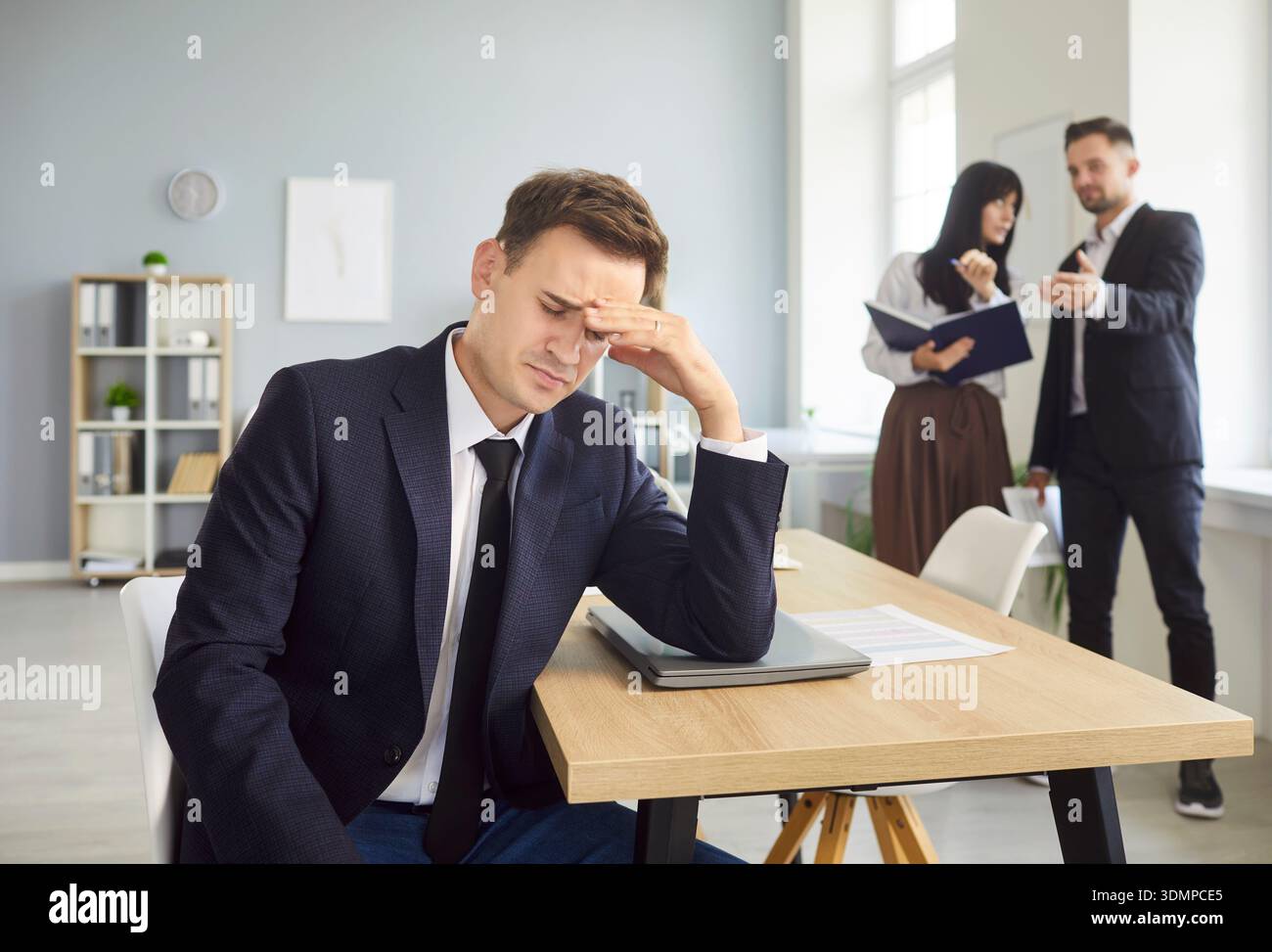 Ein frustrierter Geschäftsmann reißt sich die Stirn, während Kollegen hinter ihm über ihn sprechen Stockfoto
