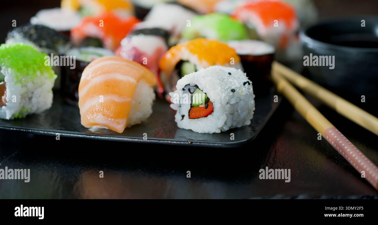 Verschiedene Sushi-Sorten, serviert auf einem schwarzen Teller mit Essstäbchen in einem japanischen Restaurant während der Mittagsstunden Stockfoto