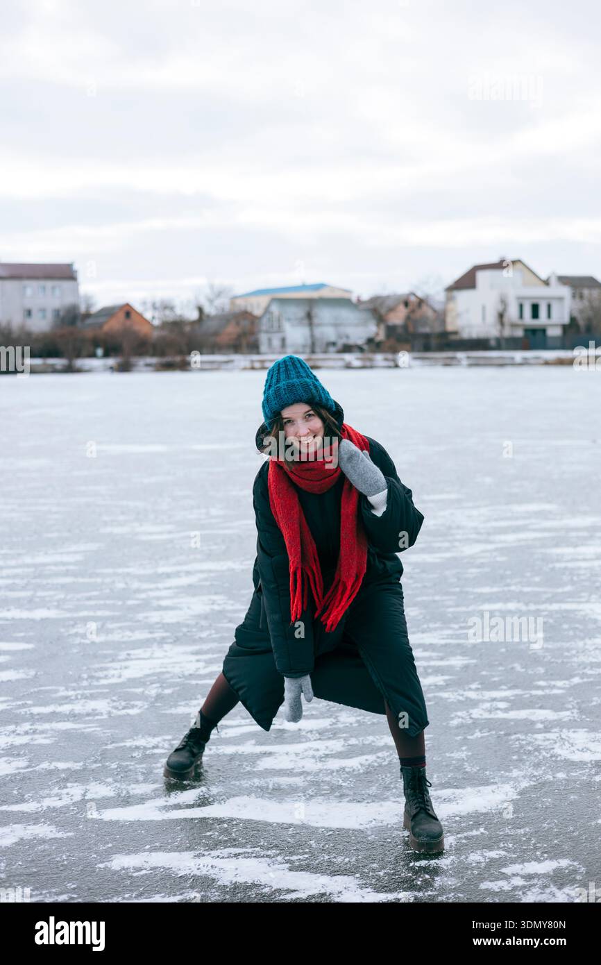 Eine Frau mit blauem Hut und rotem Schal genießt einen kalten Wintertag, wenn sie auf einem gefrorenen See steht. Häuser sind im Hintergrund sichtbar, was zu einem ruhigen Sieg führt Stockfoto
