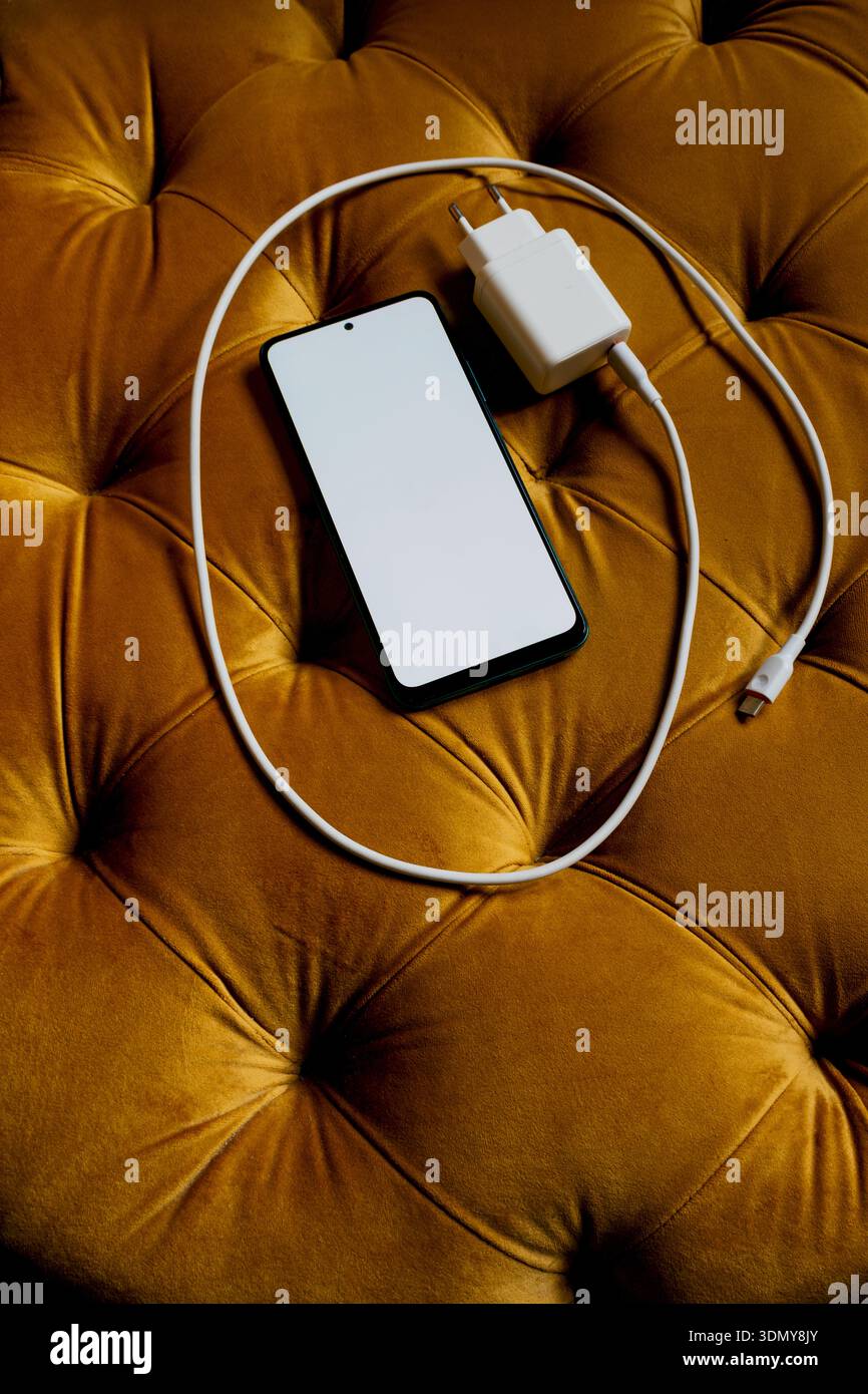 Ein modernes Smartphone mit leerem Bildschirm liegt auf einer luxuriösen Oberfläche aus Senfsamt, begleitet von einem weißen Ladekabel und Adapter, die den Text hervorheben Stockfoto