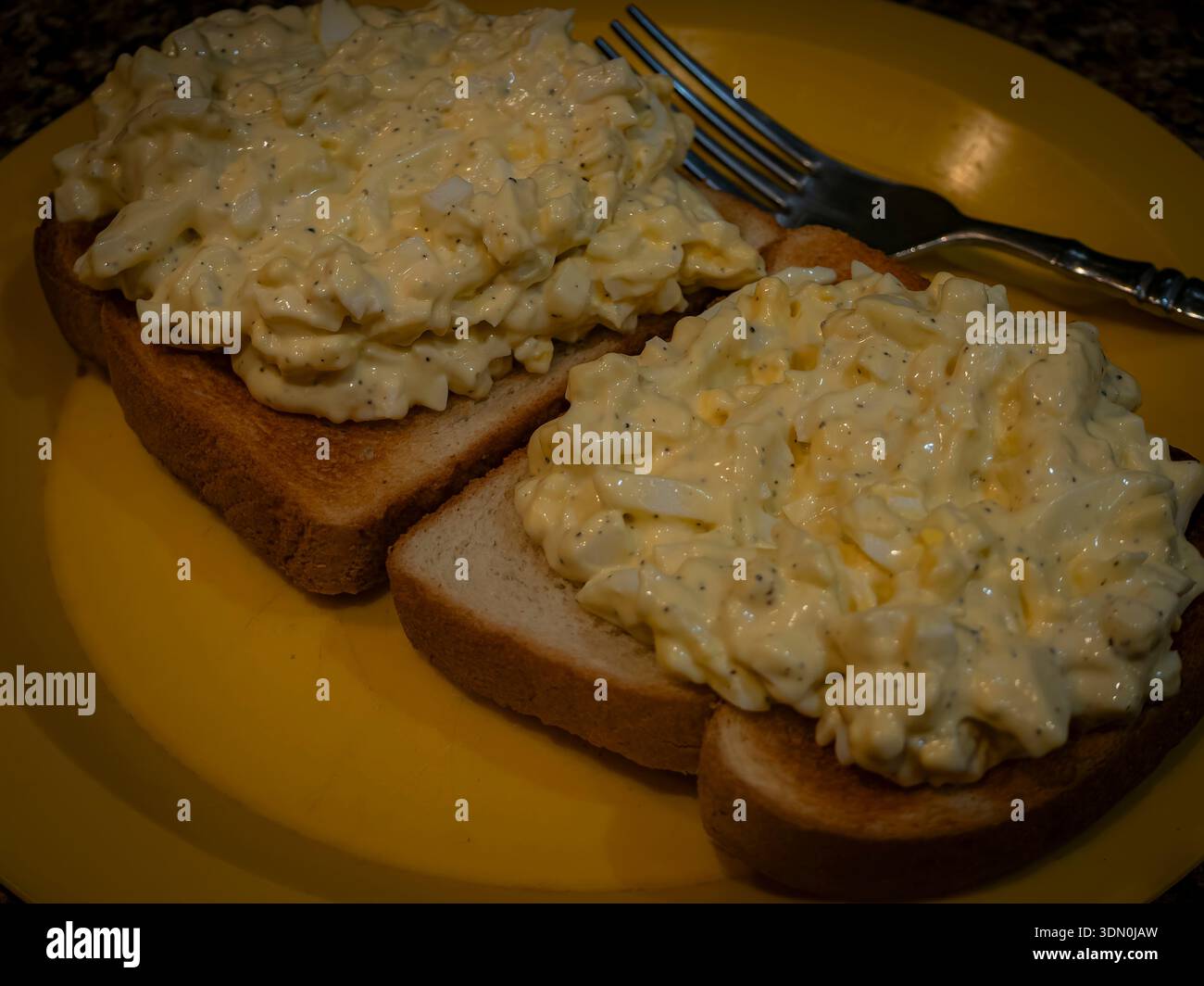 Hausgemachter Eiersalat auf geröstetem Weißbrot, serviert auf einem gelben Teller mit einer Gabel, cremige Textur und einfache, komfortable Küche. Stockfoto
