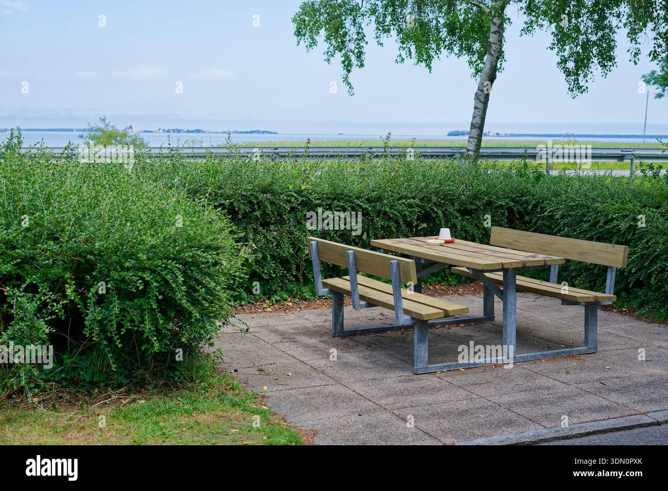 Ein kombinierter Picknicktisch und eine Sitzbank sitzt auf einem gepflasterten Einzug neben Sträuchern, hinter dem die Leitplanken sichtbar sind, wodurch eine kleine Raststätte neben dem Hi entsteht Stockfoto
