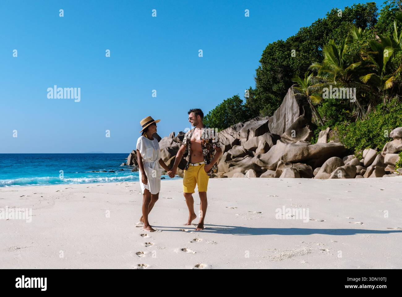 Ein fröhliches Paar geht Hand in Hand auf dem weichen weißen Sand von Anse Cocos, umgeben von üppigem Grün und kristallklarem türkisfarbenem Wasser. Die Sonne Stockfoto