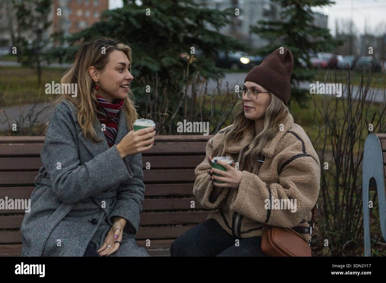 Zwei Frauen in Winterkleidung trinken Kaffee auf der Bank in der bewölkten europäischen Stadt Stockfoto