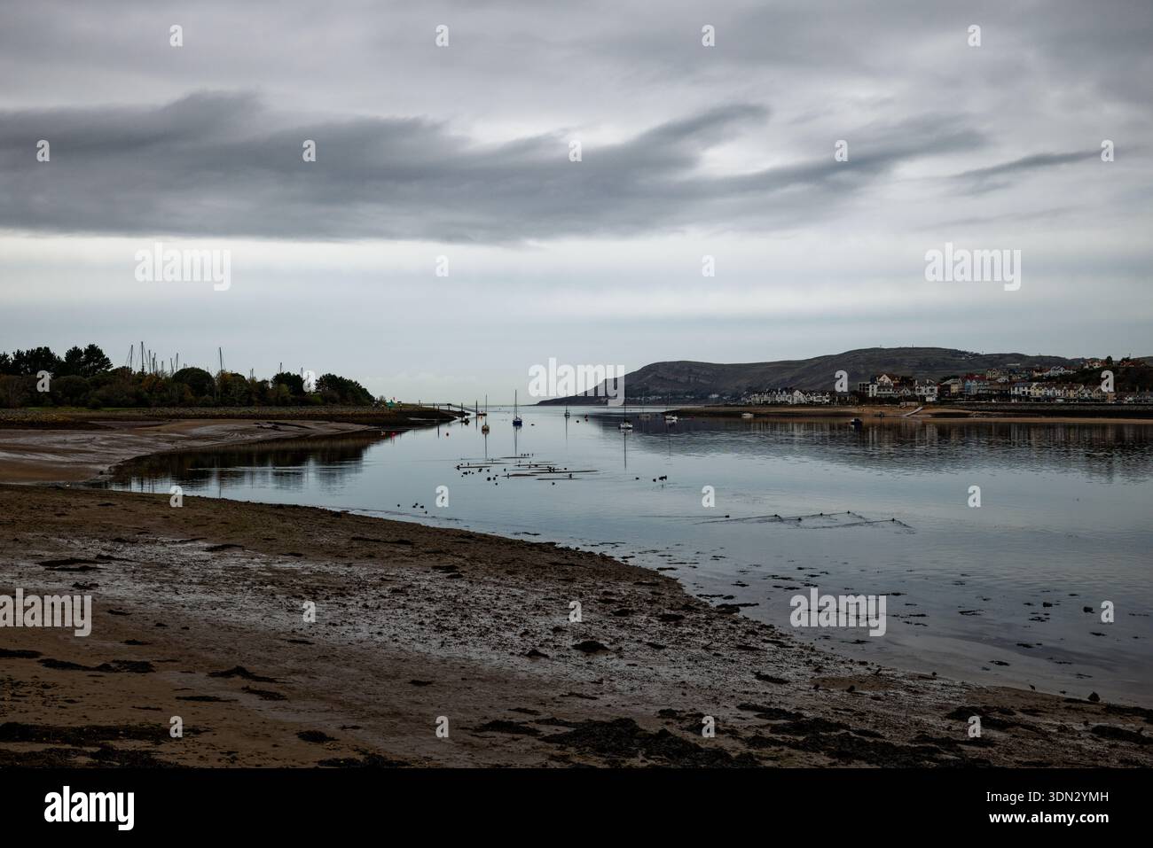 Marmorgrauer Himmel hängt bei Ebbe über den stillen Gewässern einer Mündung mit Yachten, die im mittleren Kanal in Conwy (Conway), Nordwales, ankern. Stockfoto
