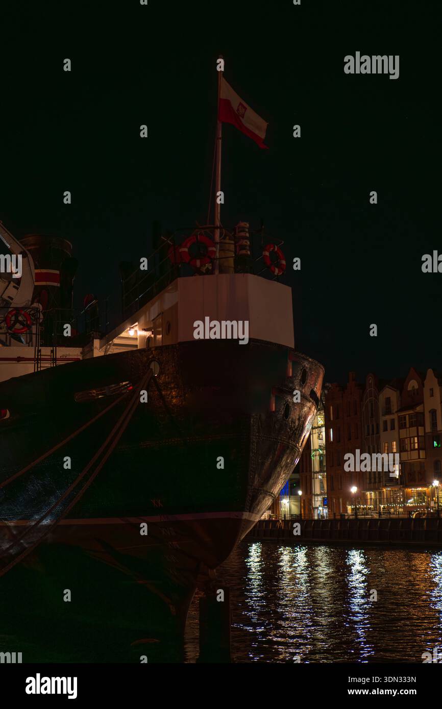 Ein historisches Schiff mit schwarzen Hüllen legt nachts auf Danzig Motlawa an. Polnische Flagge, rote und weiße Rettungsschirme, genietete Schleife und Schornstein zeichnen sich durch warme Wärme aus Stockfoto