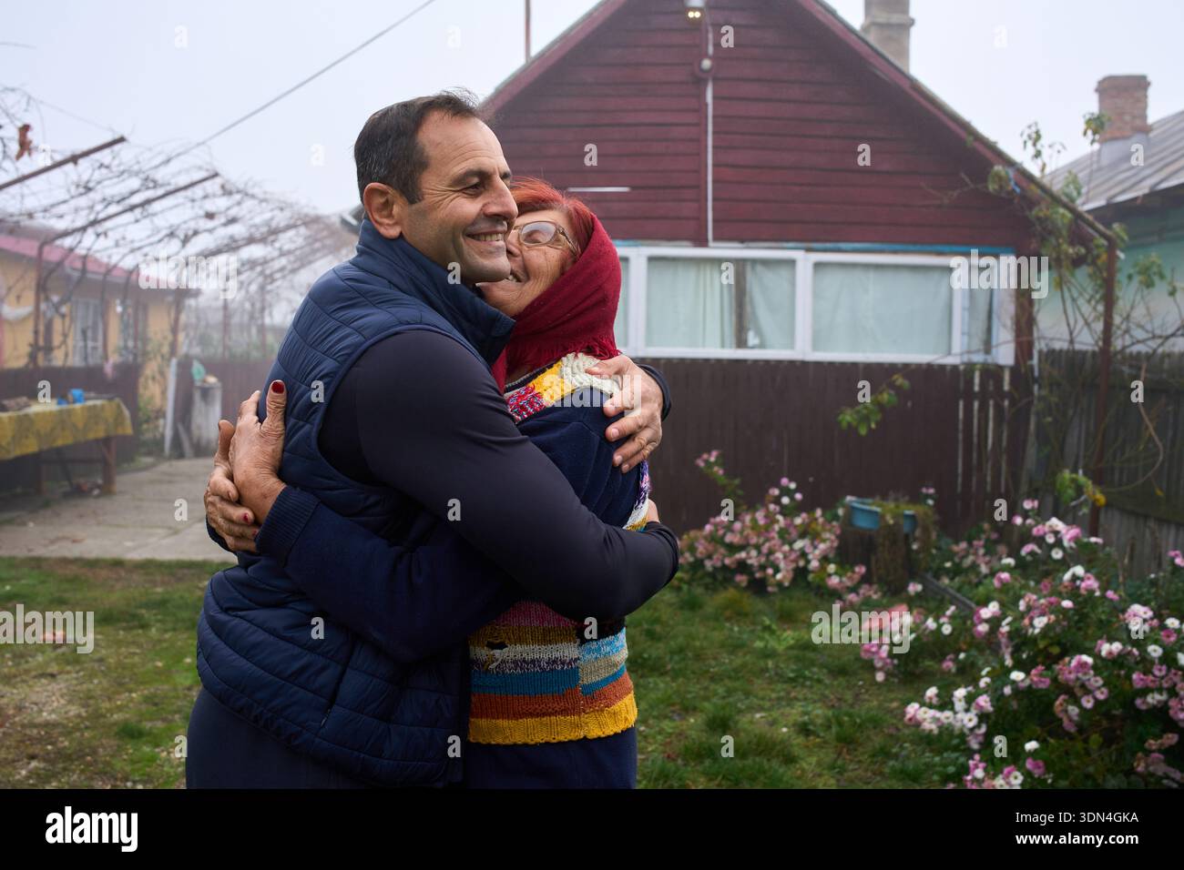 Mann mittleren Alters umarmt eine ältere Mutter vor dem ländlichen Dorfhaus Stockfoto
