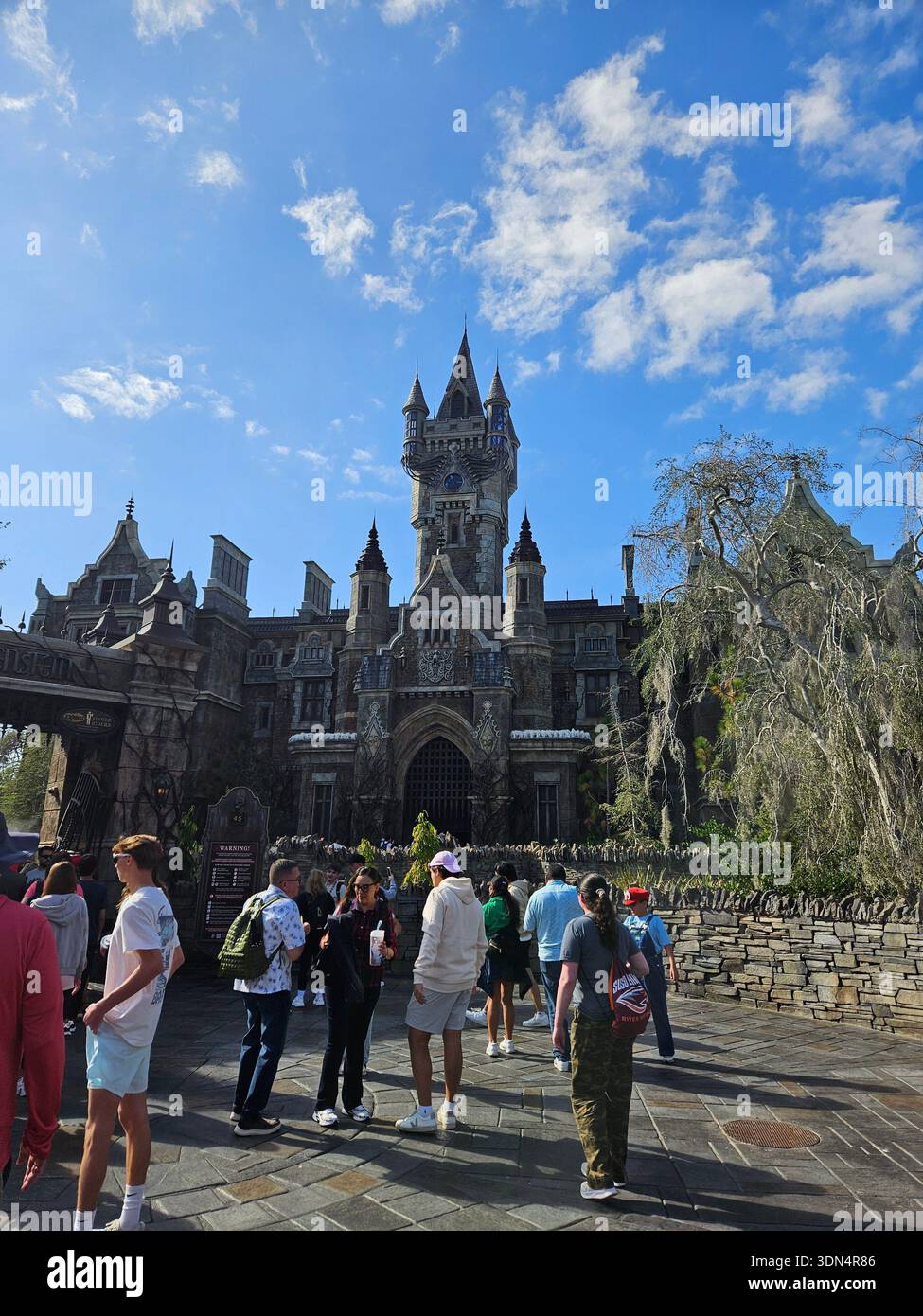 EDITORIAL USE ONLY-ORLANDO, FLORIDA-JANUAR 2026: Ein Weitwinkelblick auf das majestätische Frankenstein Manor, das Herzstück des Dunklen Universums am Universal Epic Universe. Das Bild zeigt die komplizierte, verwitterte, gotische Architektur des Herrenhauses mit einem zentralen Uhrenturm, zerklüfteten Türmen und einem großen bogenförmigen Eingang. Besucher in lässiger Kleidung werden im Vordergrund auf der kopfsteingepflasterten plaza von Darkmoor Village gesehen, die sich im Kontrast zu einem leuchtend blauen Himmel mit schimmernden Wolken befindet. Die Szene fängt die immersive, stimmungsvolle Atmosphäre des Themenbereichs Universal Monsters an einem hellen Tag ein. Stockfoto