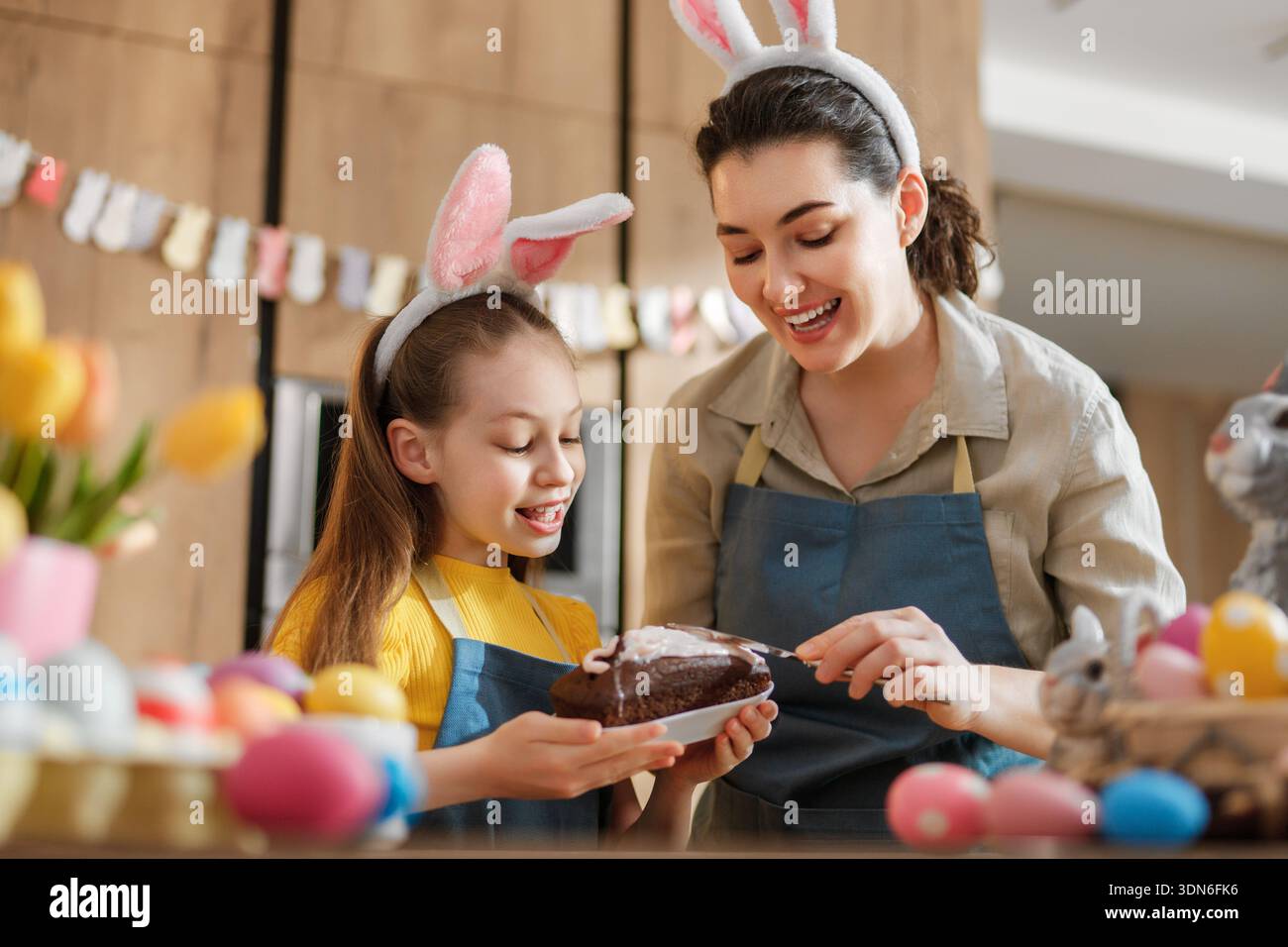 Fröhliche Familienbindung in der Küche, während Mutter und Tochter Schokoladenkuchen schmücken. In der Ostertradition tragen sie verspielte Hasenohren Stockfoto