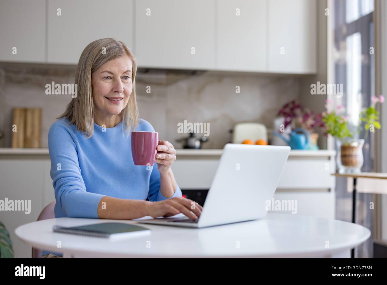 Eine ältere Frau lächelt und tippt auf einem Laptop, hält einen Becher und arbeitet von zu Hause aus an einem weißen Tisch in ihrer modernen Küche Stockfoto
