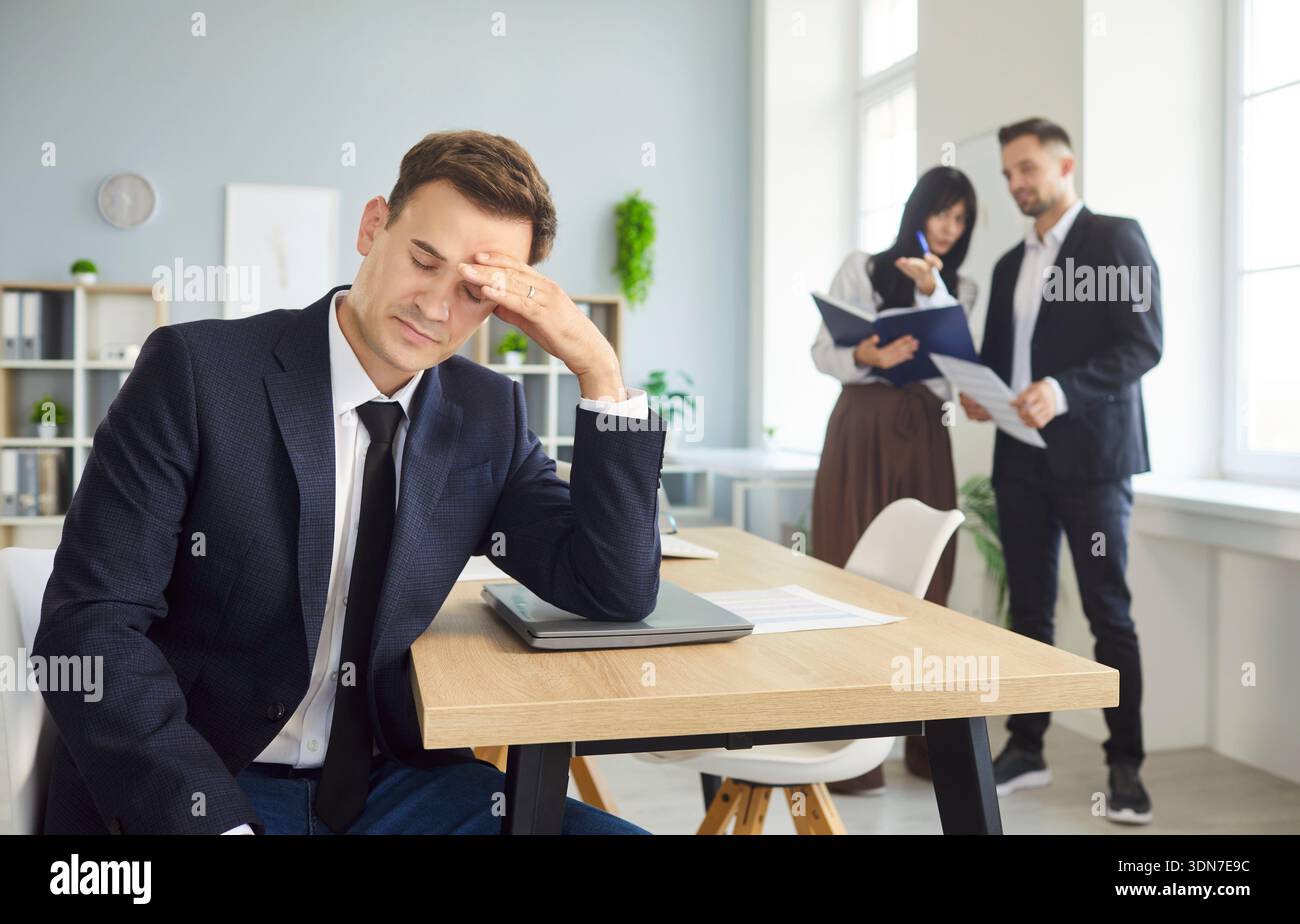 Gestresster isolierter männlicher Mitarbeiter am Schreibtisch, während Kollegen im Hintergrund sprechen Stockfoto