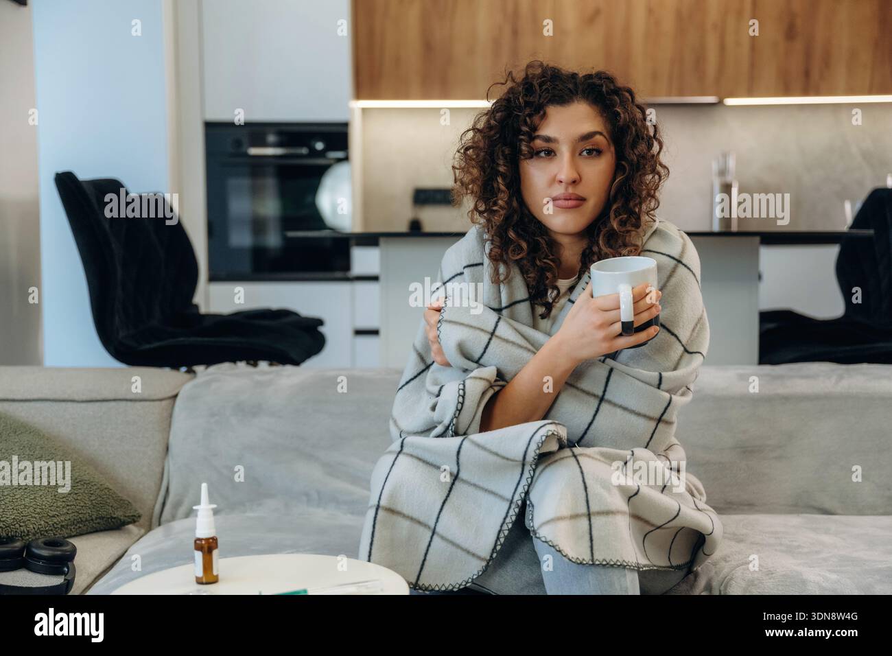 Der weiße Becher liegt in der Hand. Kranke Frau, die sich zu Hause unwohl fühlt, ihre Temperatur überprüft und Medikamente einnimmt, während sie sich auf einem Sofa ausruht. Stockfoto