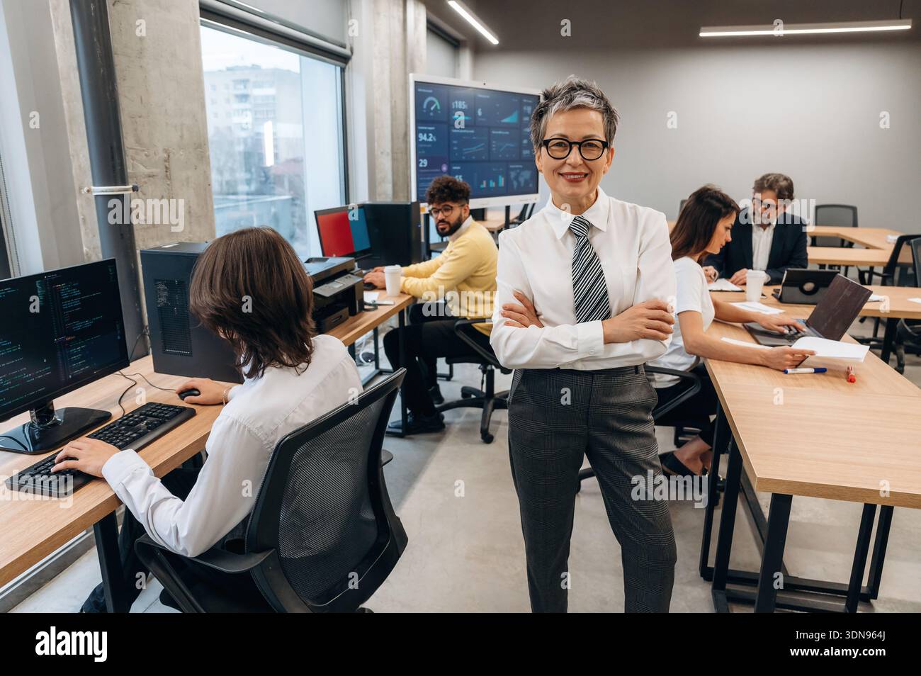 Geschäftsfrau mit Brille und grauem Haar steht selbstbewusst in einem modernen Büro, während Kollegen mit Computern arbeiten und Teamwork und Arbeit hervorheben Stockfoto