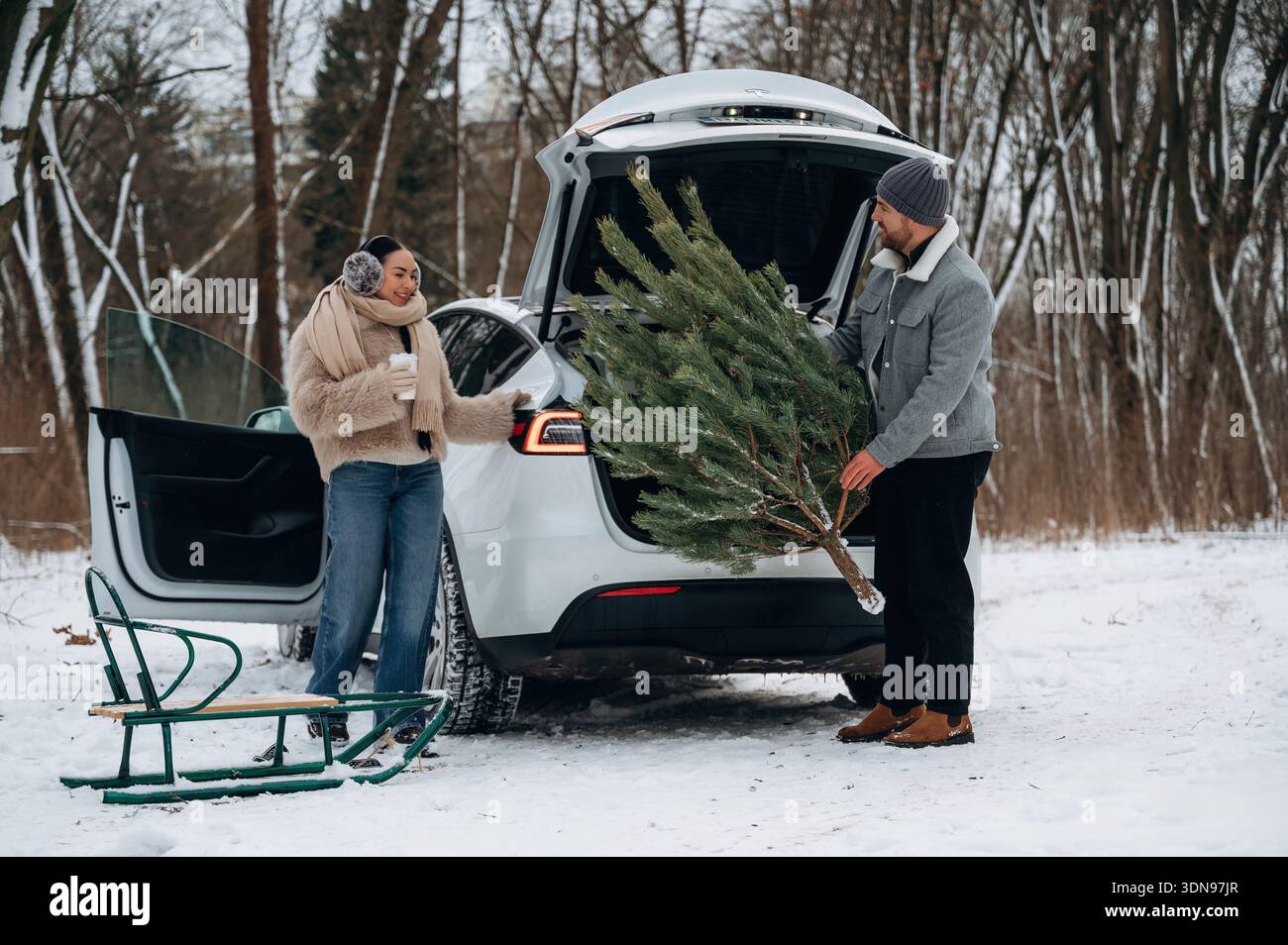 Ein junges Paar, eine Frau in einem gemütlichen Pullover und ein Mann in einer warmen Jacke, die einen Weihnachtsbaum in den Kofferraum in einer verschneiten Umgebung lädt, was einen fröhlichen Urlaub in Erinnerung ruft Stockfoto