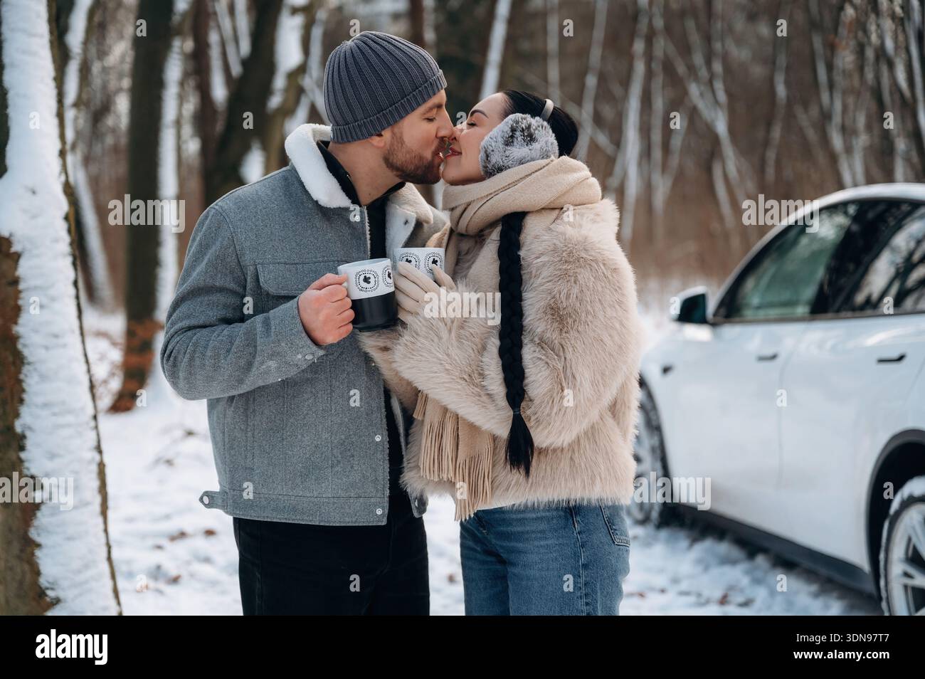Ein Paar, in gemütlicher Winterkleidung gekleidet, teilt sich einen Kuss, während es in einem verschneiten Wald, umgeben von Bäumen, Tassen hält, was einen warmen und intimen geldautomaten schafft Stockfoto