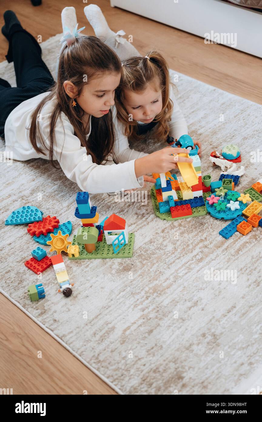 Zwei Mädchen liegen auf einem weichen Teppich und konzentrieren sich darauf, farbenfrohe Strukturen mit Spielzeugblöcken zu bauen, um Kreativität und Teamarbeit in einer gemütlichen Innenumgebung zu fördern Stockfoto