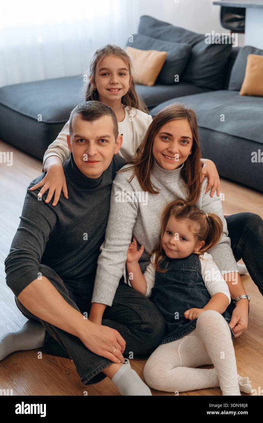 Vierköpfige Familie, einschließlich Eltern und zwei Töchtern, sitzt zusammen auf einem gemütlichen Wohnzimmerboden, umgeben von weichen Möbeln und natürlichem Licht Stockfoto