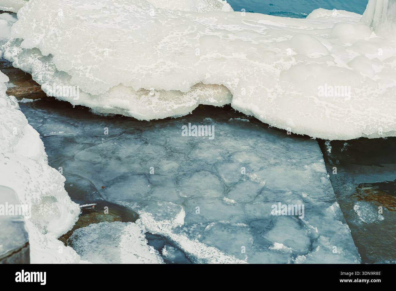 In einer Winterszene bedecken Eis und Schnee die Oberfläche eines Gewässers. Blaue Wasserflecken sind im Eis sichtbar, was auf ein kaltes Klima hinweist. Stockfoto