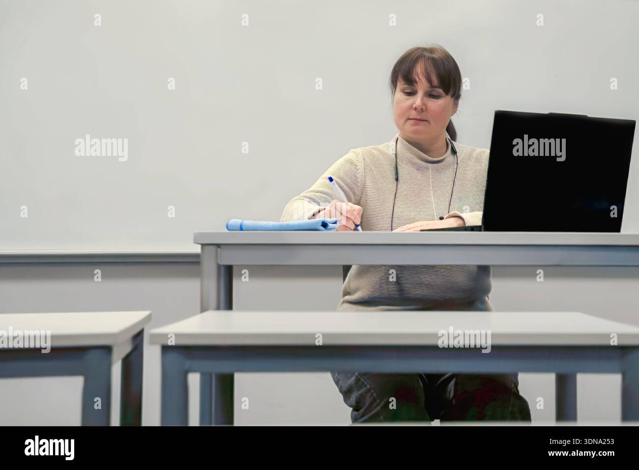 Frau mit braunem Haar sitzt an einem Schreibtisch, schreibt Notizen und benutzt einen Laptop in einem hellen Klassenzimmer, um Konzentration und lehrreiche Atmosphäre zu betonen Stockfoto