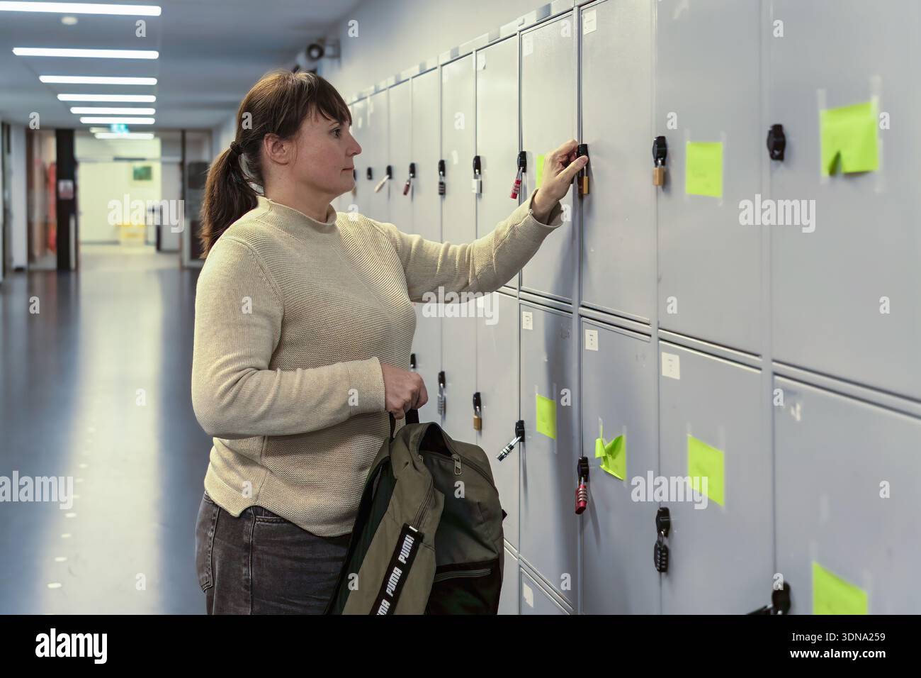 Weibliche Person in beigefarbenem Pullover kommt in einem gut beleuchteten Flur zu einem Schließfach, umgeben von grauen Schließfächern, was die persönliche Organisation und dai betont Stockfoto