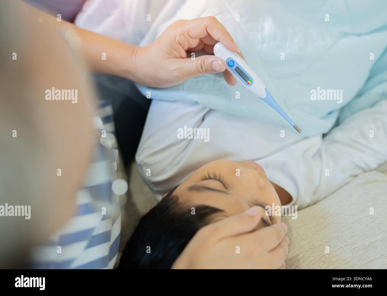 Hochwinkelansicht eines im Bett liegenden asiatischen Kranken mit einem Thermometer in der Hand seiner Mutter. Krankes Kind Konzept. Stockfoto