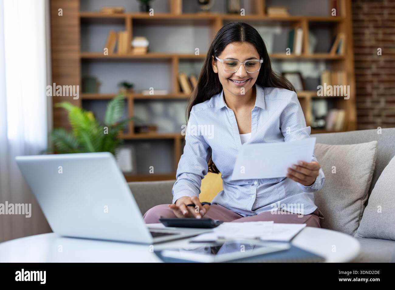 Junge Frau lächelt, während sie zu Hause sitzt, Dokumente sorgfältig überprüft, Taschenrechner und Laptop benutzt, persönliche Finanzen verwaltet und Steuern vorbereitet Stockfoto