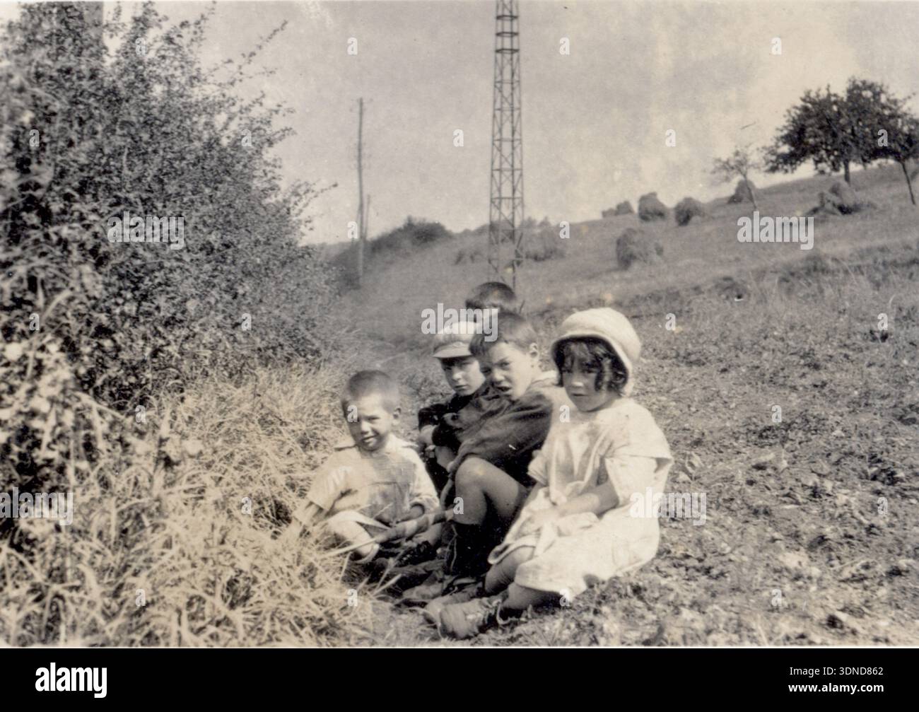 Fünf Kinder sitzen auf dem Gras neben einem Busch und überblicken schräge Felder mit Heuhaufen. In der Ferne erhebt sich ein hoher Metallturm, der sich von der ländlichen Landschaft abhebt. Ihre Kleidung und die pastorale Umgebung halten einen ruhigen 1927-Moment nahe Elvreux fest. Aus einer Sammlung einzigartiger Fotografien des 20. Jahrhunderts. Amateurfotograf, Schnappschüsse und einige größere in einem einzigen Album, das die Reisen einer Familie in Europa dokumentiert. Stockfoto