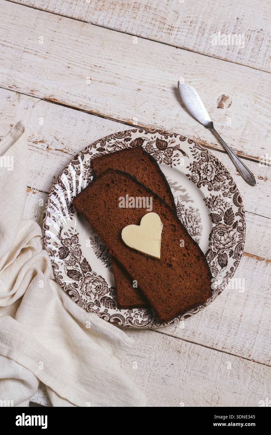 Eine Liebesbotschaft zum Frühstück. Ein Stück herzförmige Butter auf einem dunklen Roggenbrot. Ansicht von oben nach unten Stockfoto